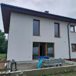 Dwukondygnacyjny dom po tynkowaniu i malowaniu elewacji na biało, widoczne drewniane ramy okienne, teren wokół domu w trakcie porządkowania, częściowo zakryty niebieską plandeką.