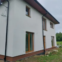 Dwukondygnacyjny dom z jasną elewacją, brązowymi oknami i orynnowaniem, widok z boku na tle pochmurnego nieba i zieleni.