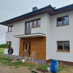 Usługi budowlane, Tomasz Wochnik, Henryk Wochnik - Dwupiętrowy dom z białą elewacją i drewnianymi oknami, z fragmentem drewnianej fasady nad wejściem, w trakcie prac wykończeniowych; widoczne drabiny, puszki z farbą i pracownik w ubrudzonych farbą...