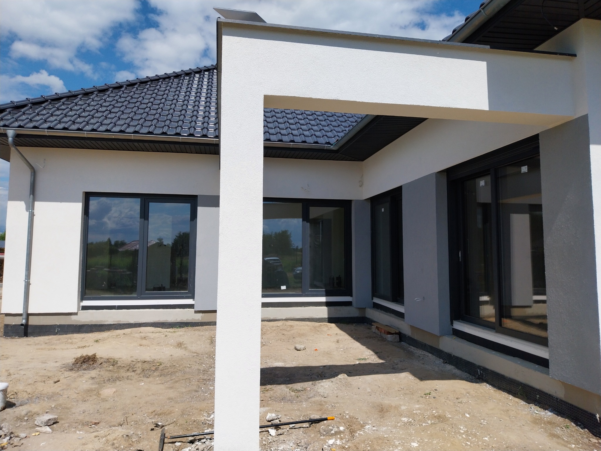 Nowoczesny dom jednorodzinny z szarą elewacją i dużymi oknami w czarnych ramach, widoczny fragment otynkowanej ściany z elementami wykończeniowymi przy gruncie. Teren wokół domu jeszcze...