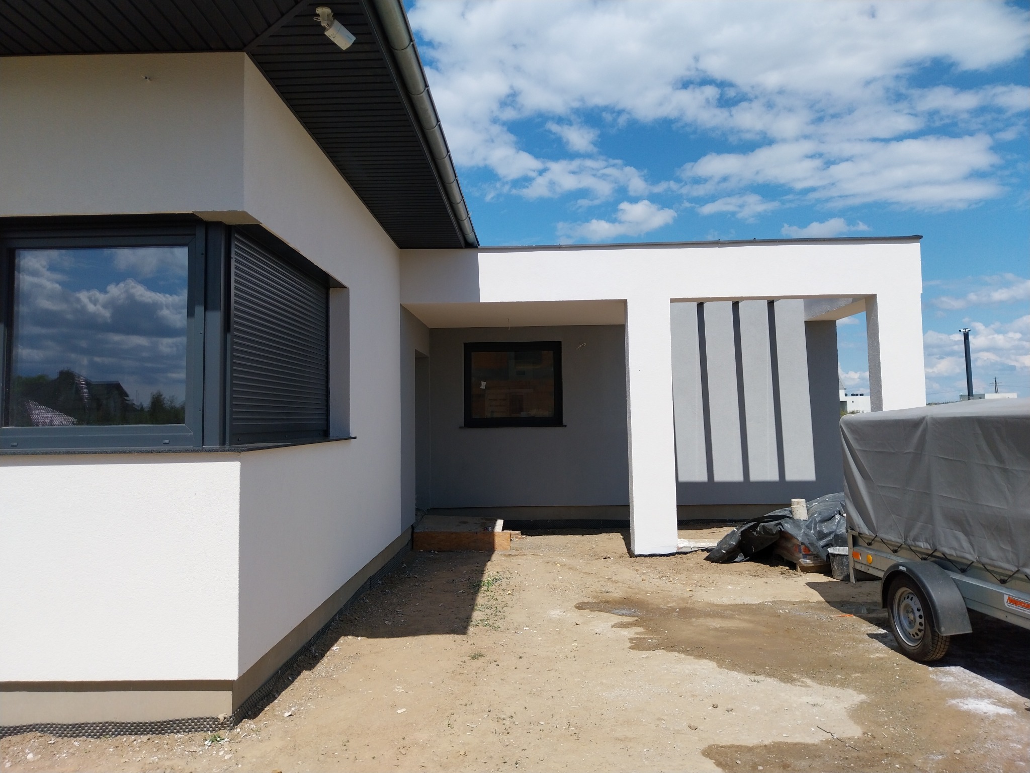 Nowoczesny dom jednorodzinny z jasną elewacją i ciemnymi oknami, widoczny fragment elewacji z roletami zewnętrznymi, fragment podjazdu z przyczepką, błękitne niebo z chmurami odbija się w szybach...