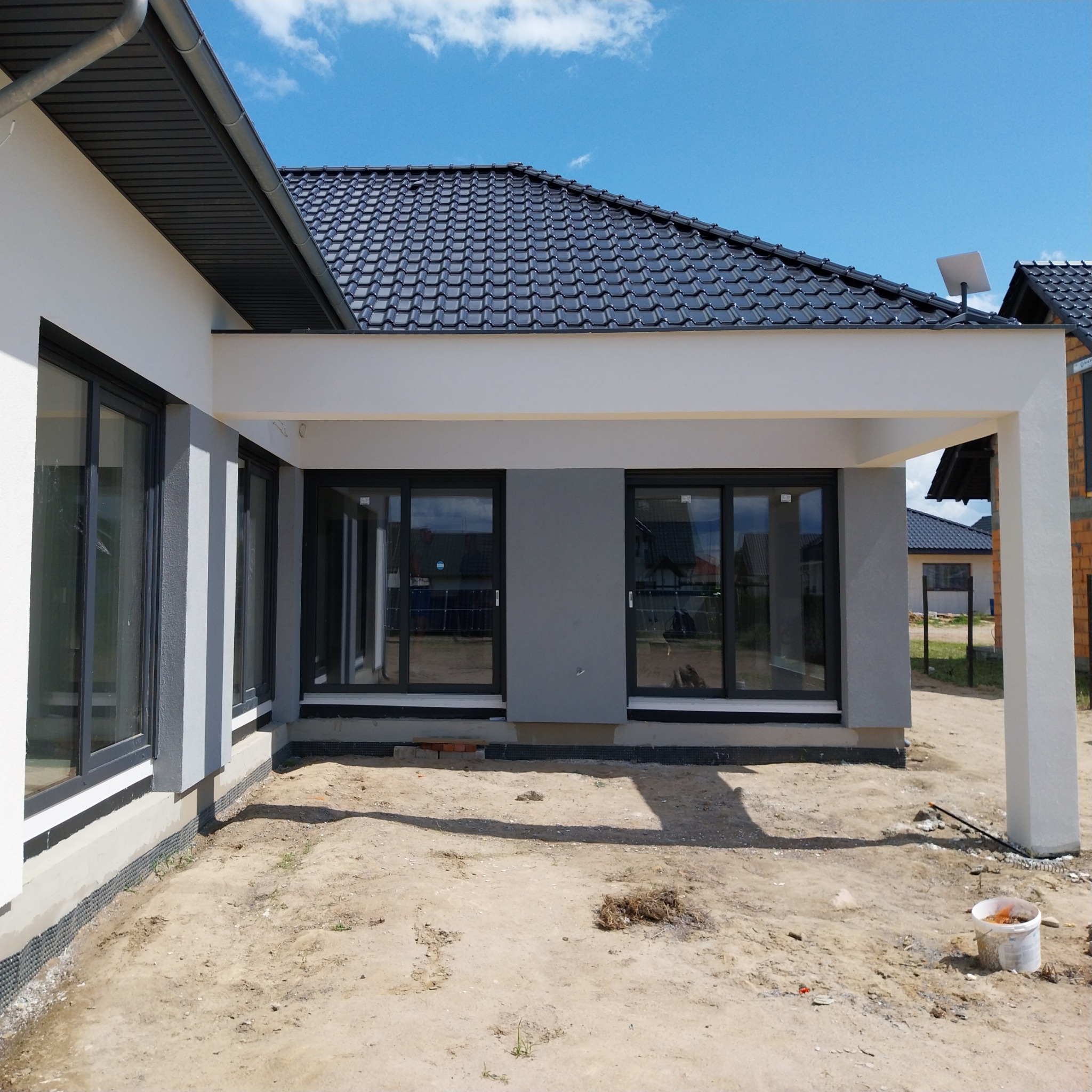 Nowoczesny dom jednorodzinny z szarą elewacją i czarnymi oknami, widok z boku na zewnątrz, teren wokół domu przygotowany pod zagospodarowanie, widoczne ślady prac budowlanych i wiadro po farbie.