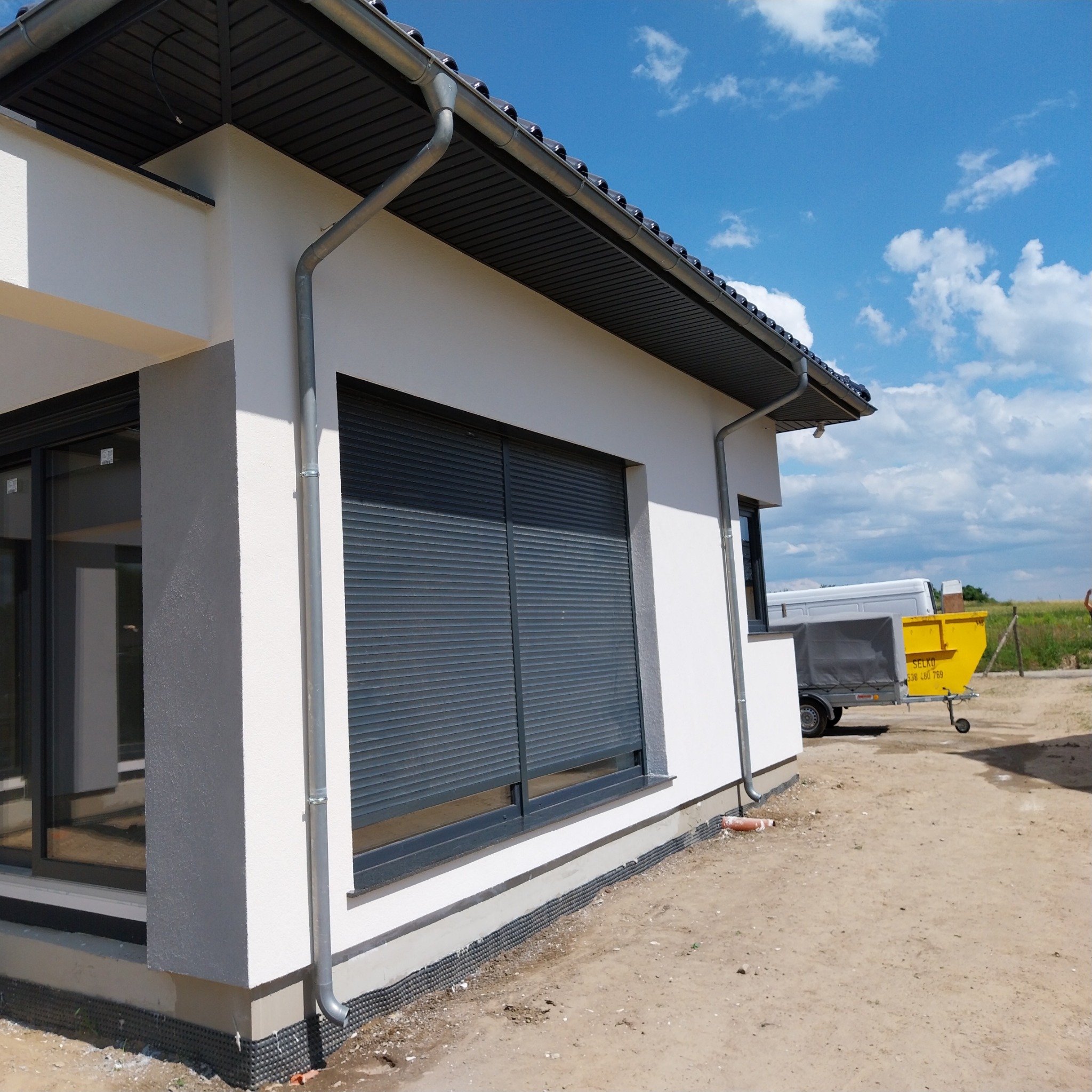 Fragment elewacji domu jednorodzinnego z szarymi roletami zewnętrznymi, widoczne rynny, biały tynk i fragment podsypki pod elewację oraz przyczepka z kontenerem w tle.