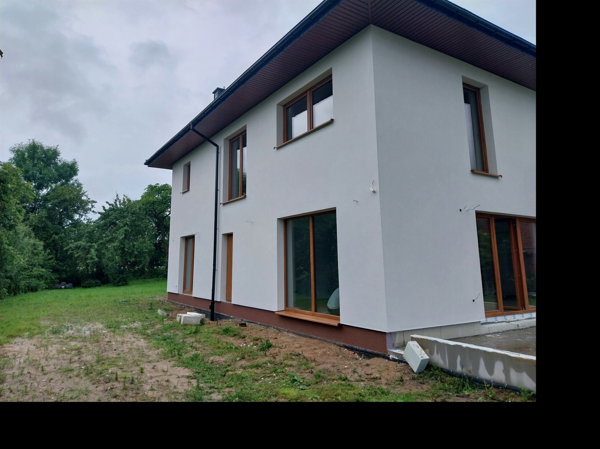 Dwupiętrowy dom z białą elewacją i brązowymi ramami okiennymi, widoczny z zewnątrz na tle zieleni, z fragmentami niezakończonych prac wykończeniowych wokół fundamentów i niezagospodarowanym terenem.
