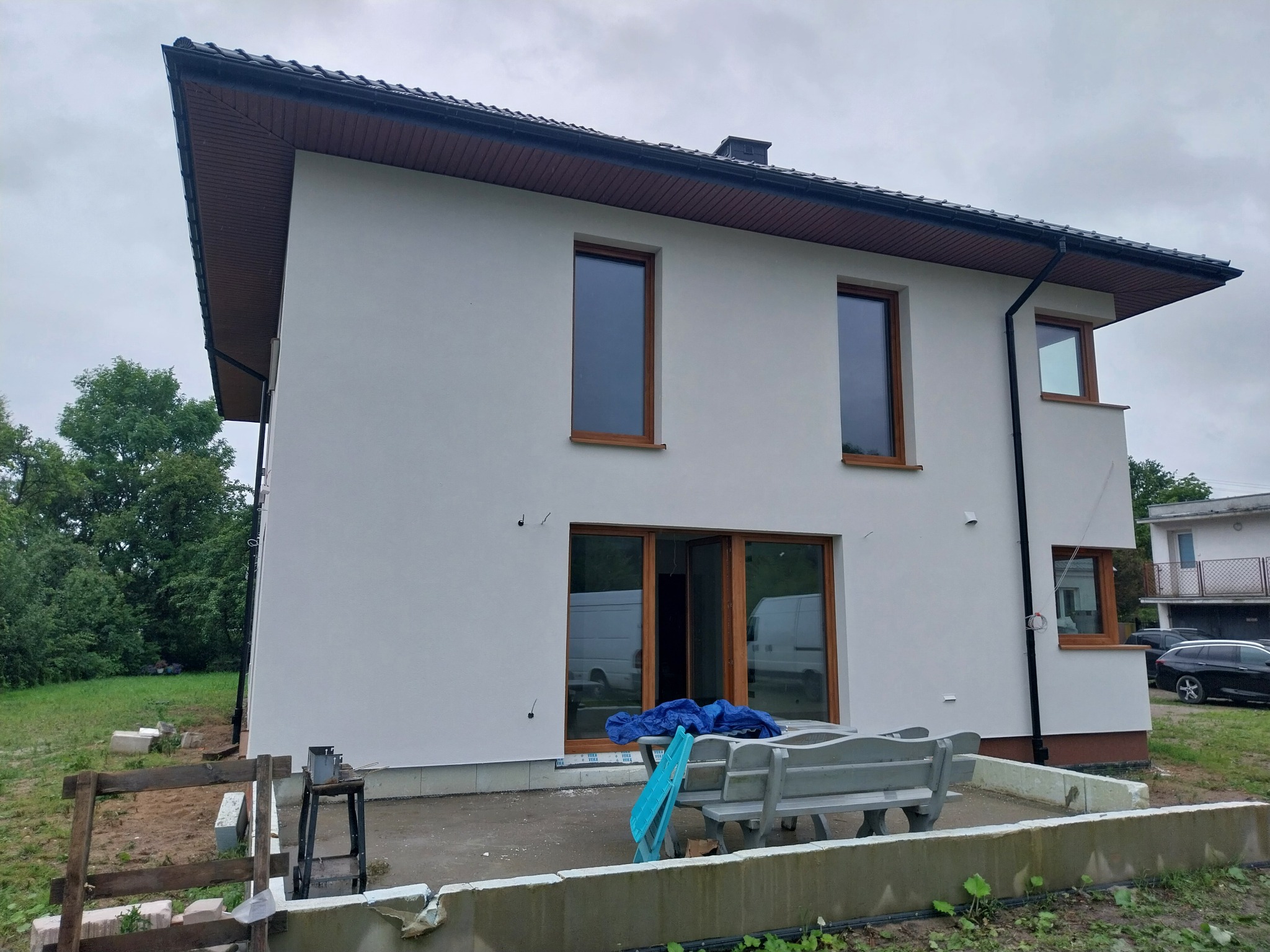 Dwukondygnacyjny dom po tynkowaniu i malowaniu elewacji na biało, widoczne drewniane ramy okienne, teren wokół domu w trakcie porządkowania, częściowo zakryty niebieską plandeką.