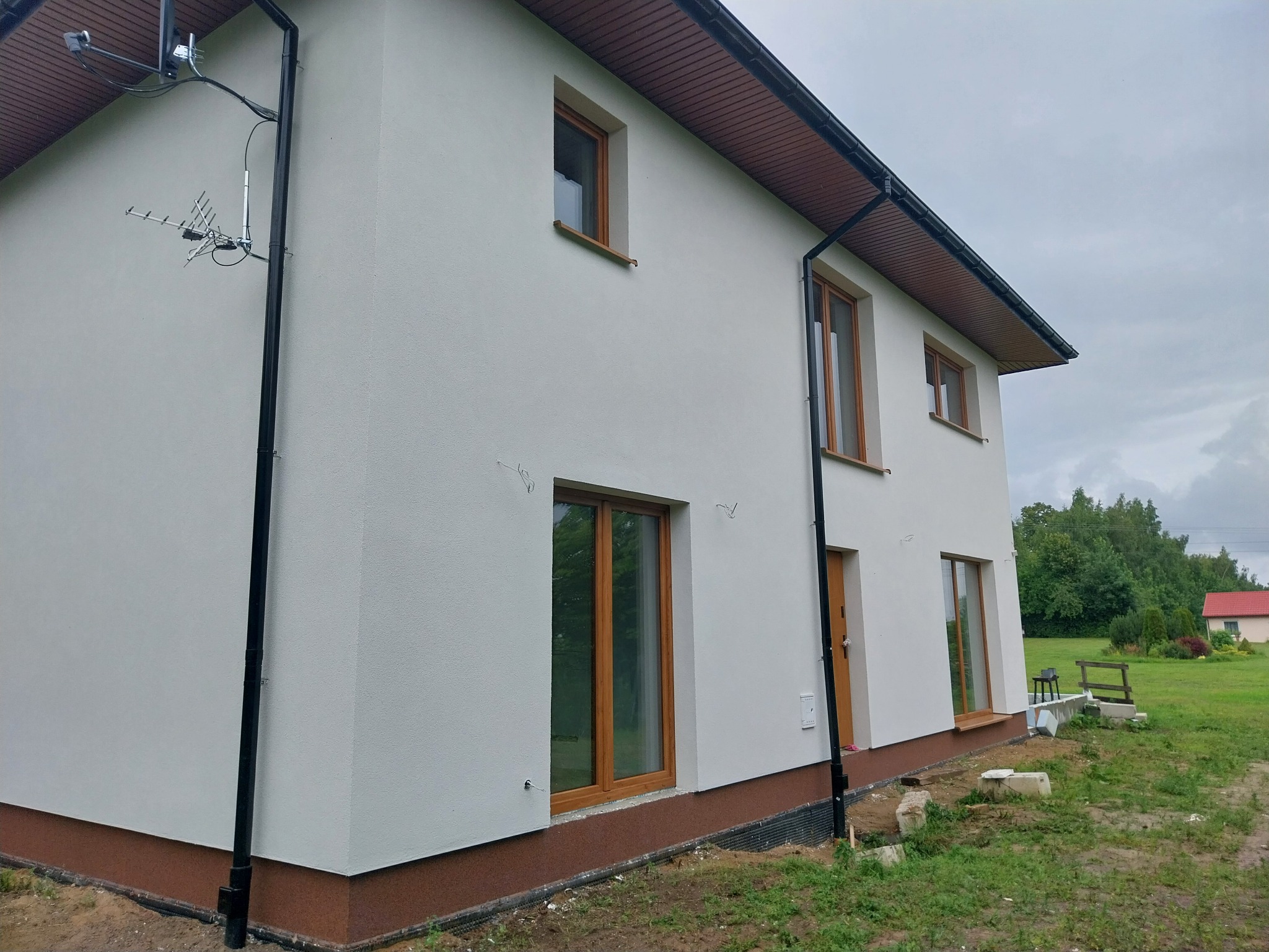 Dwukondygnacyjny dom z jasną elewacją, brązowymi oknami i orynnowaniem, widok z boku na tle pochmurnego nieba i zieleni.