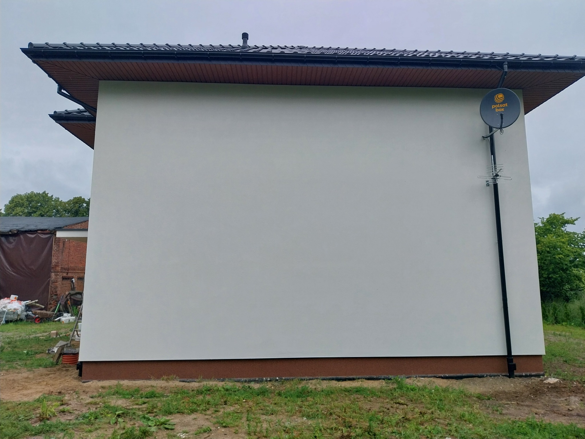 Świeżo otynkowana ściana budynku z zamontowaną anteną satelitarną Polsat Box, widoczne ślady prac budowlanych w tle.