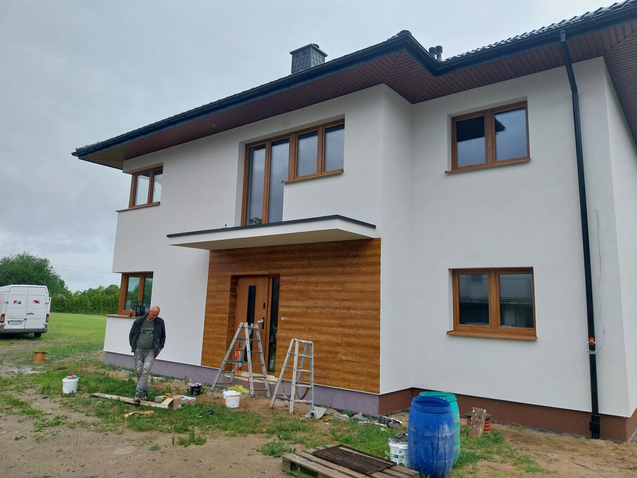 Dwupiętrowy dom z białą elewacją i drewnianymi oknami, z fragmentem drewnianej fasady nad wejściem, w trakcie prac wykończeniowych; widoczne drabiny, puszki z farbą i pracownik w ubrudzonych farbą...