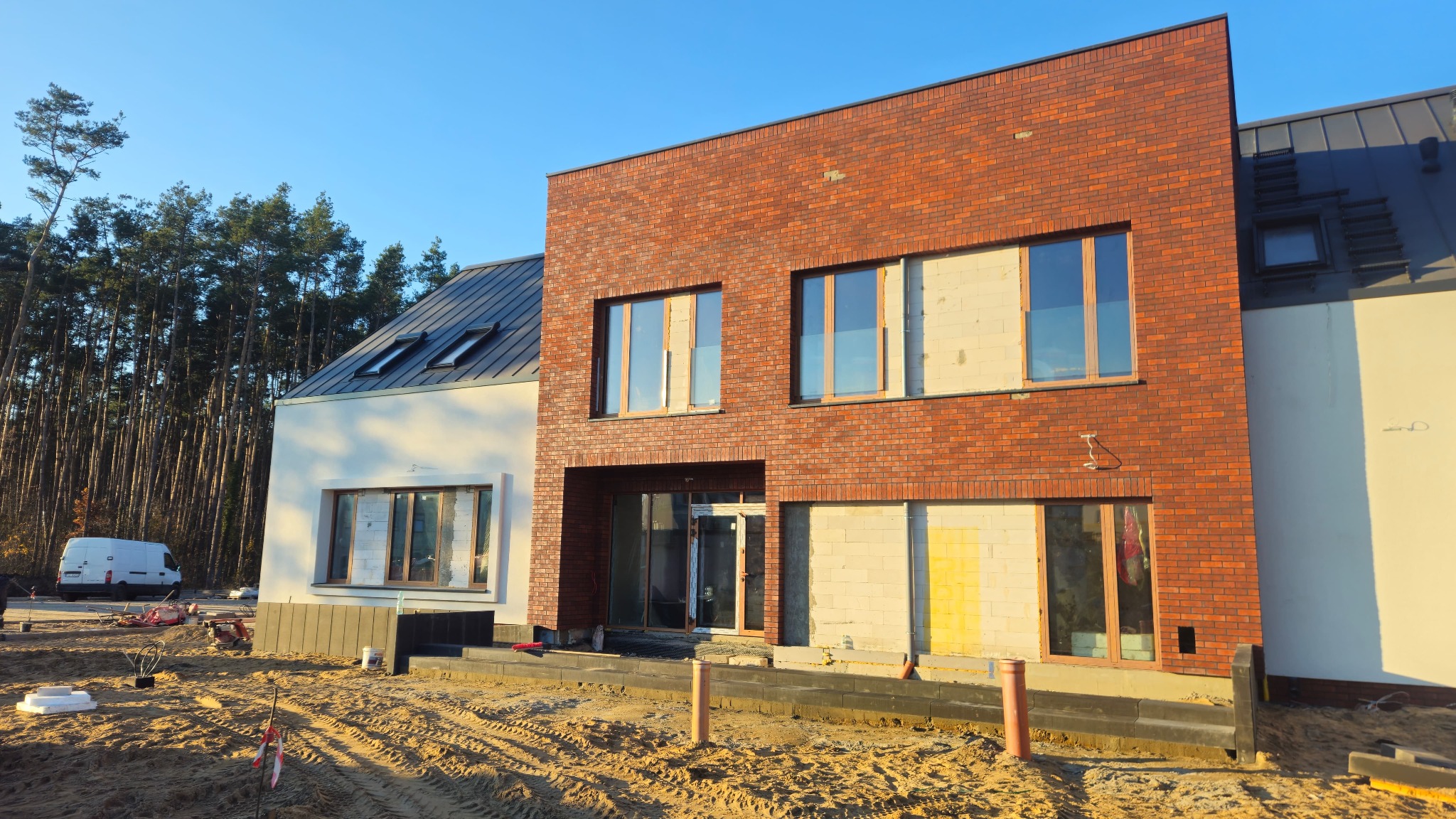 Dwukondygnacyjny dom z ceglaną elewacją w trakcie budowy, widoczne niezabezpieczone okna i elementy konstrukcyjne. W tle las i błękitne niebo.