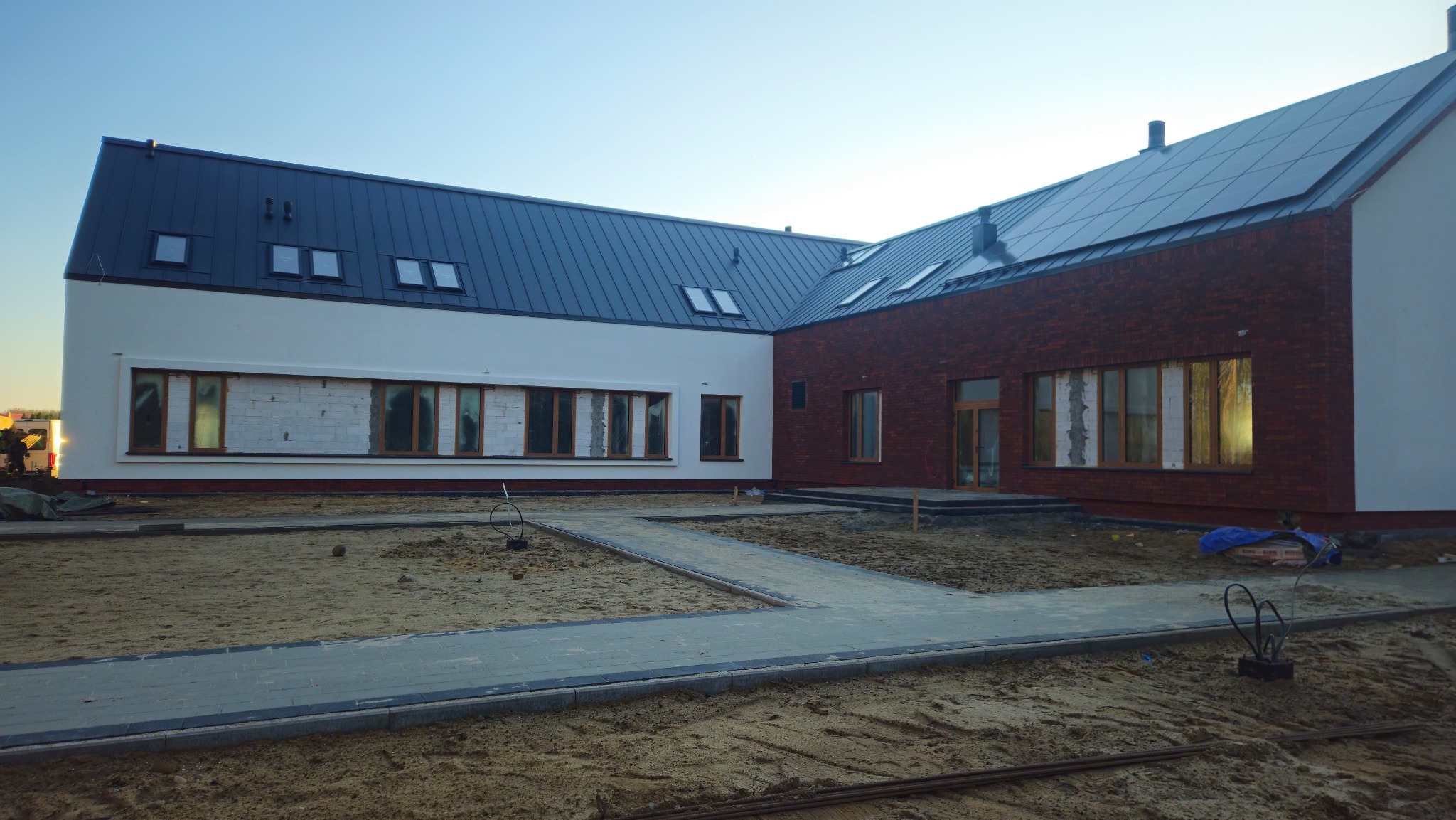 Nowoczesny budynek z białą i ceglaną elewacją pod ciemnym dachem. Widoczne okna z drewnianymi ramami i częściowo otynkowana ściana. Teren wokół w trakcie prac wykończeniowych.