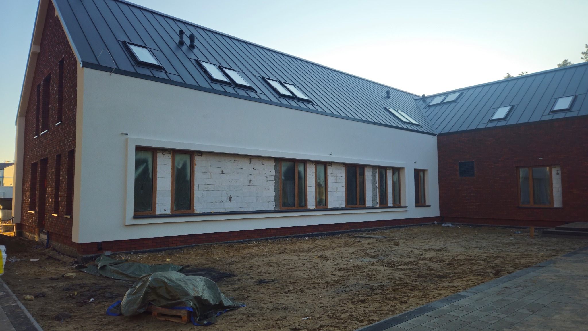 Nowoczesny dom z białymi ścianami i ceglanymi elementami. Widoczne okna, część ściany w trakcie prac wykończeniowych. Dach z oknami dachowymi.