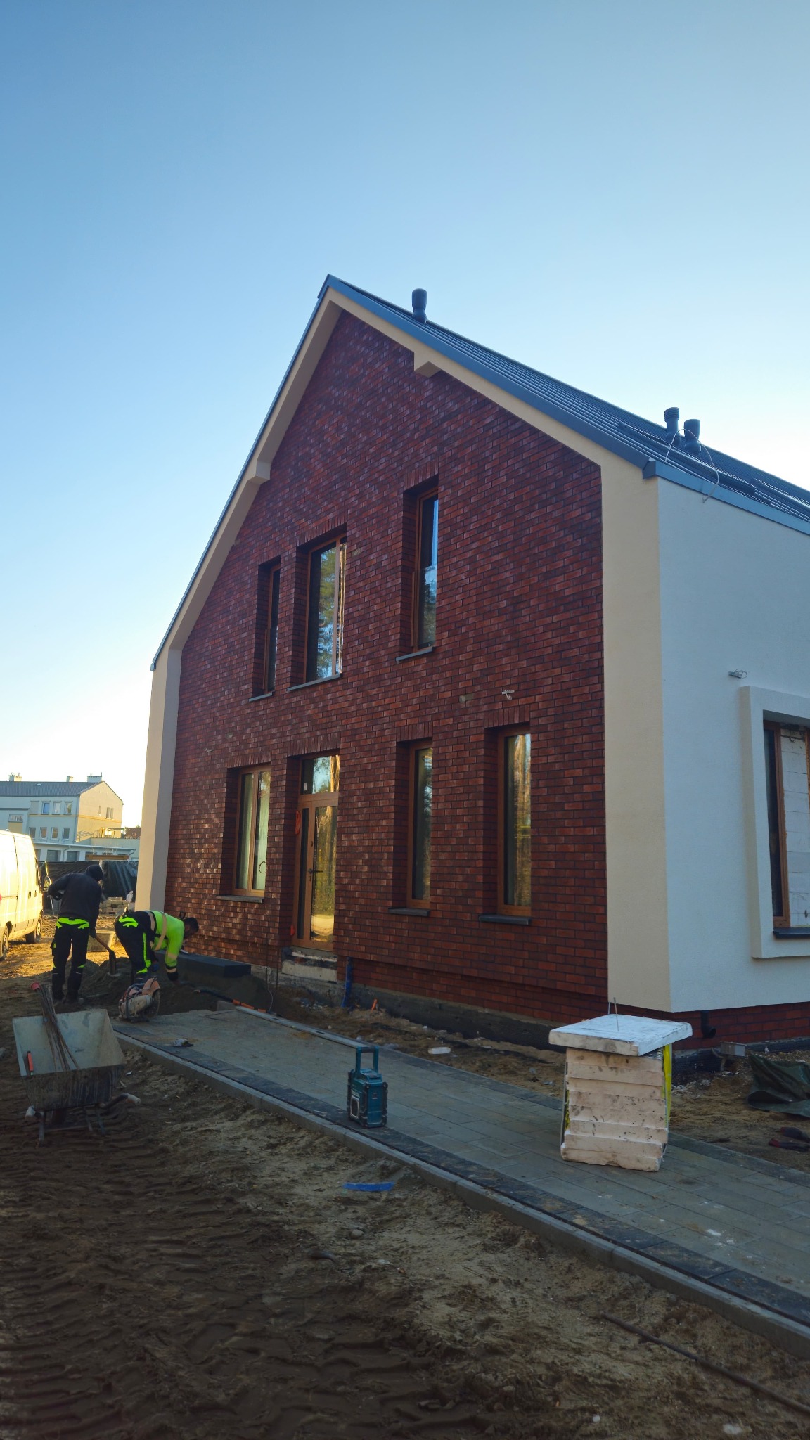 Dwukondygnacyjny dom z ceglaną elewacją i białym tynkiem, widoczni pracownicy w trakcie prac wykończeniowych na zewnątrz budynku, w słoneczny dzień.
