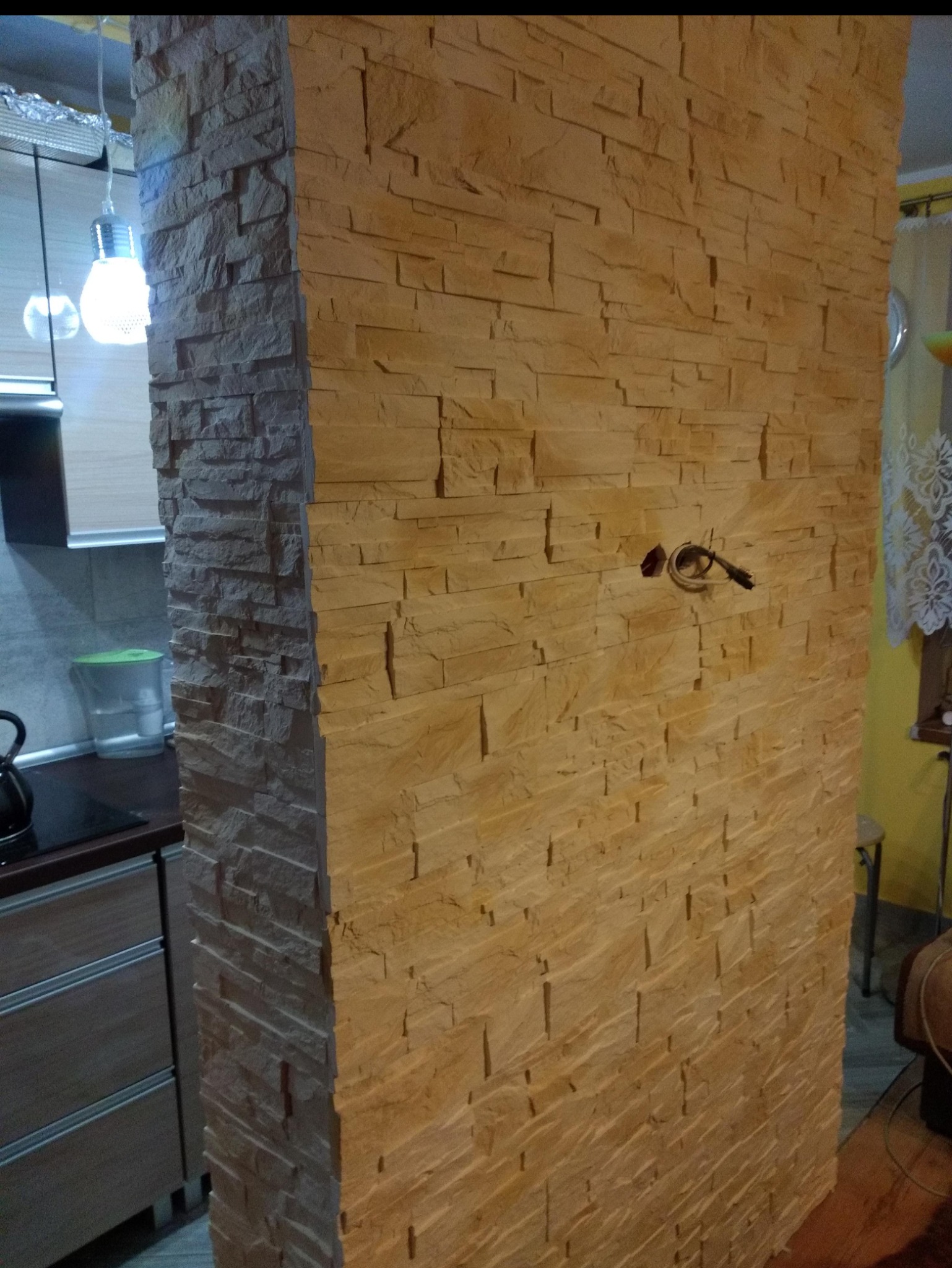 Ściana wykończona dekoracyjnym kamieniem w odcieniach beżu i brązu, z widocznym wystającym przewodem elektrycznym, fragment kuchni z oświetleniem w tle.