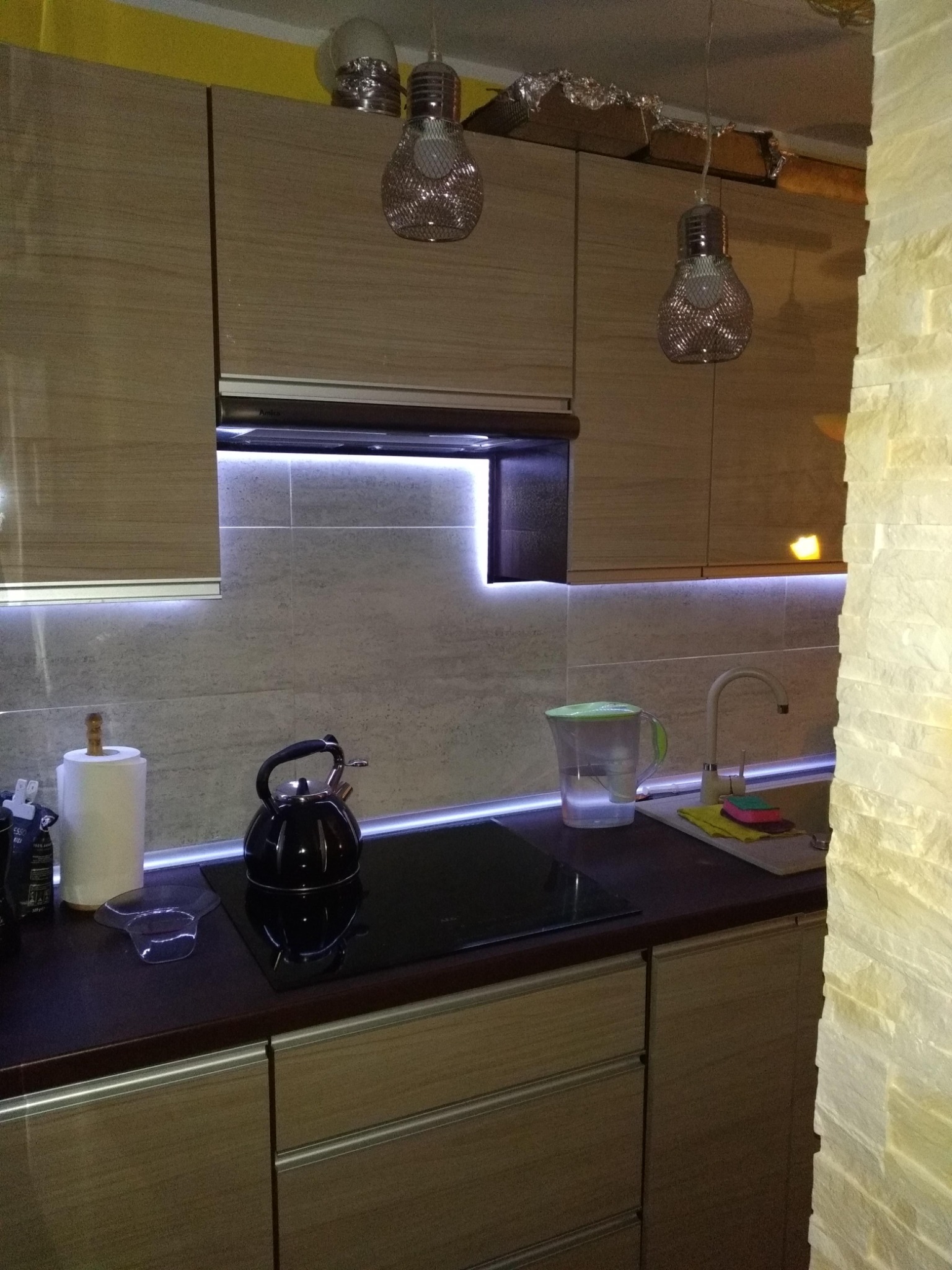 Nowoczesna kuchnia z drewnianymi szafkami, podświetleniem LED pod szafkami wiszącymi i dekoracyjnymi lampami wiszącymi w kształcie żarówek. Na blacie czajnik, dzbanek filtrujący i akcesoria kuchenne.