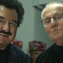 Portret dwóch mężczyzn, jeden z wąsami i słuchawką w uchu, uśmiechnięty, drugi w okularach, obaj ubrani w ciemne ubrania na rozmytym tle.