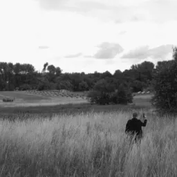 Czarno-białe ujęcie mężczyzny w garniturze, idącego przez pole z wysoką trawą, w tle widoczne czołgi, przeszkody przeciwpancerne i samoloty na niebie, sugerujące scenę filmową lub rekonstrukcję...