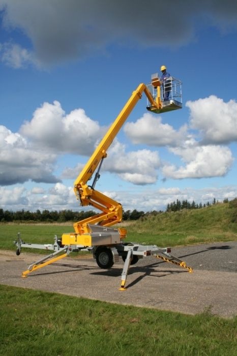 Żółty podnośnik koszowy na przyczepie z pracownikiem w koszu na tle błękitnego nieba z chmurami, ustawiony na asfalcie obok trawnika.