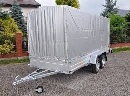 Aluminiowa przyczepa dwuosiowa z plandeką w kolorze srebrnym, zaparkowana na żwirowym podłożu, z fragmentem ceglanego ogrodzenia i zielenią w tle.