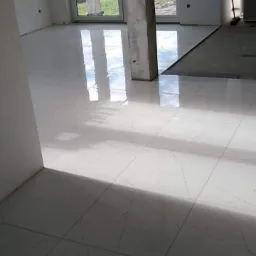 Błyszcząca, biała podłoga z płytek imitujących marmur, świeżo ułożona w jasnym pomieszczeniu z dużym oknem i surowym filarem, widoczne krzyżyki dystansowe.