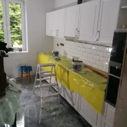 Wnętrze kuchni w trakcie remontu: białe szafki wiszące, białe płytki na ścianie nad blatem zabezpieczonym żółtą folią, drabina aluminiowa, podłoga wyłożona płytkami imitującymi marmur.