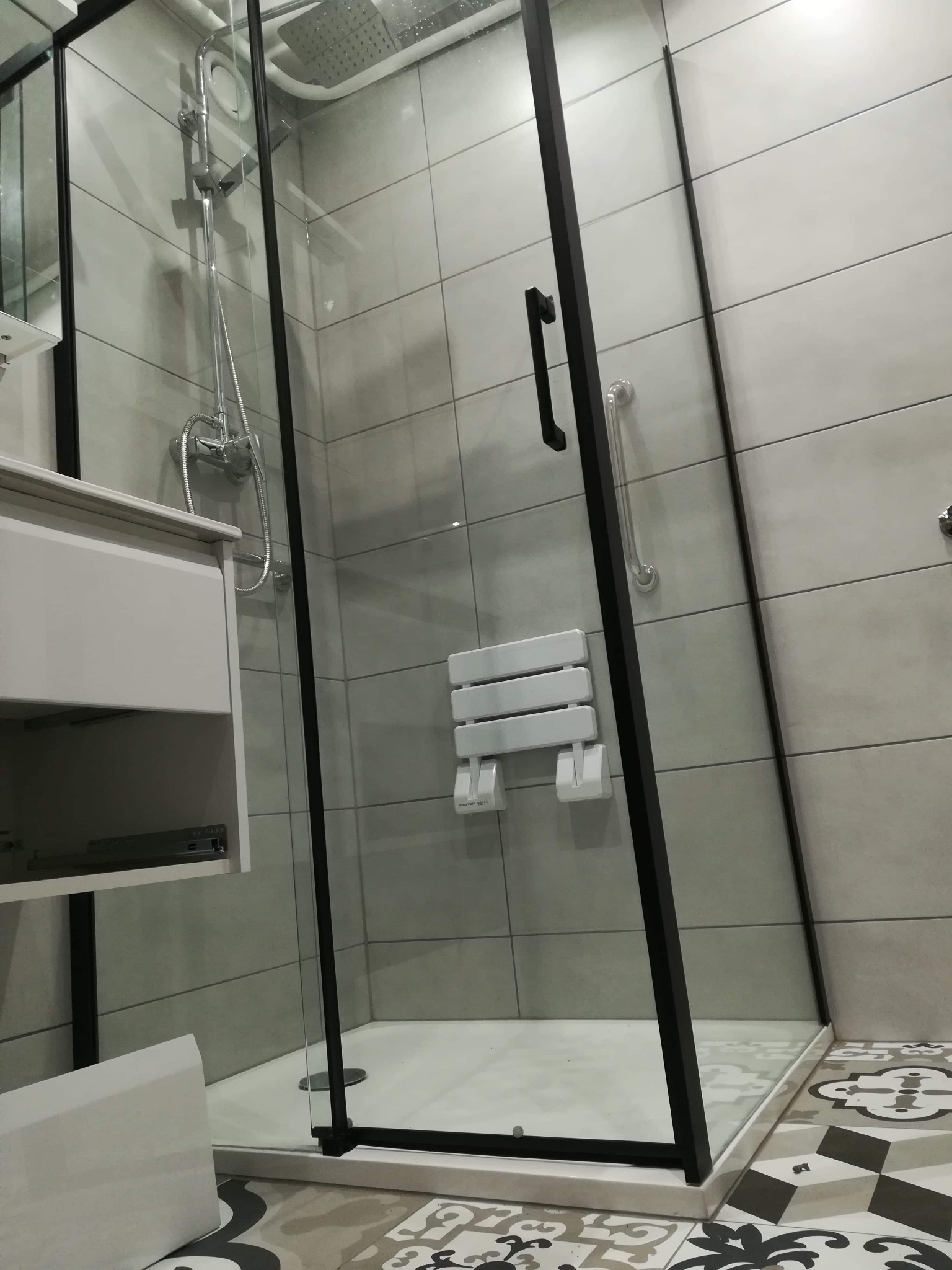 Nowoczesna kabina prysznicowa z czarnymi profilami, szarymi płytkami i siedziskiem, widoczna umywalka z szafką i wzorzysta podłoga.