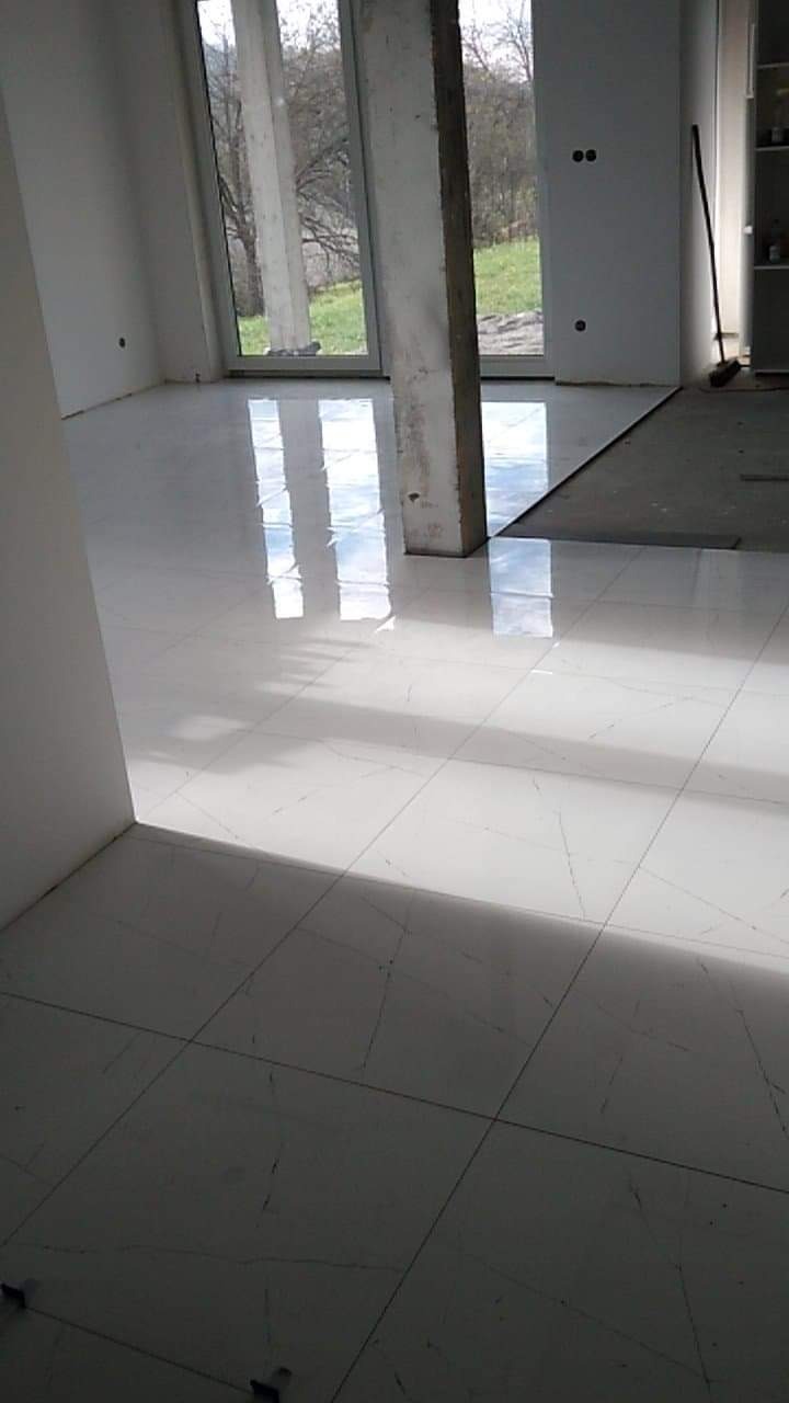 Błyszcząca, biała podłoga z płytek imitujących marmur, świeżo ułożona w jasnym pomieszczeniu z dużym oknem i surowym filarem, widoczne krzyżyki dystansowe.