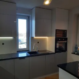 Nowoczesna kuchnia z szarymi frontami, czarnym blatem i oświetleniem LED pod szafkami wiszącymi, z piekarnikiem i mikrofalówką w zabudowie, widok na okno z zewnętrznym krajobrazem.