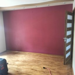 T.P Usługi Tomasz Piszczek - Ściana pomalowana na bordowo w pokoju z drewnianą podłogą i otwartymi drzwiami. Widoczny odkurzacz przemysłowy i kabel zasilający.