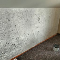 T.P Usługi Tomasz Piszczek - Teksturowana, biała ściana po szpachlowaniu w skosie poddasza, zabezpieczona folią i z wiaderkiem farby. Widok na wykończone wnętrze.