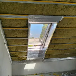Ocieplenie poddasza z oknem dachowym: wełna mineralna między metalowymi profilami, zabezpieczona folią aluminiową. Widok na zewnątrz przez okno.