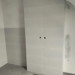 Jasne, prostokątne płytki ścienne i podłogowe w łazience, widoczne otwory na armaturę i odpływ podłogowy. Minimalistyczny design z szarymi fugami.