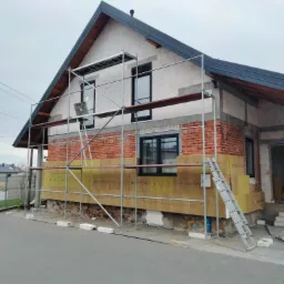 Dom w trakcie ocieplania elewacji, widoczne rusztowanie, częściowo ocieplona ściana z cegły i bloczków, żółta wełna mineralna, drabina oparta o rusztowanie.