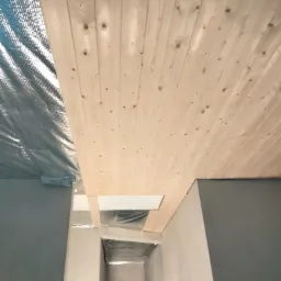 Montaż drewnianej boazerii na suficie z widoczną izolacją termiczną, surowe drewno z sękami, fragment szarej ściany i widok na korytarz w tle.