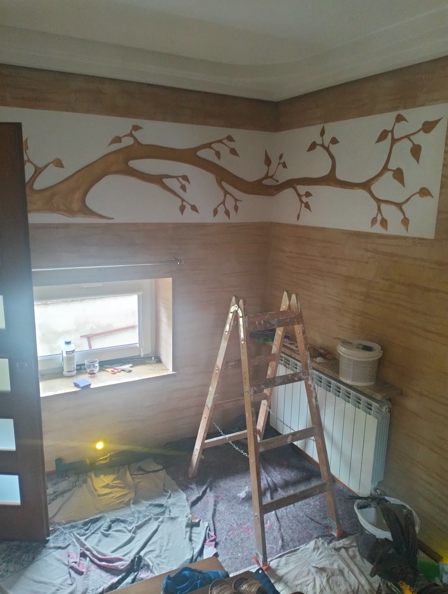 Pokój po dekoracyjnym wykończeniu ścian z motywem drzewa. Widoczna drabina malarska, okno z akcesoriami i zabezpieczona podłoga. Ciepła kolorystyka.