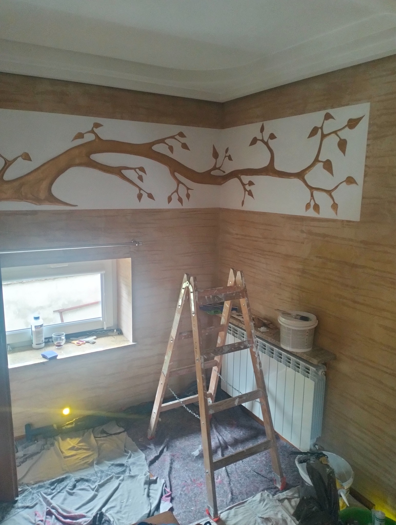 Wykończone wnętrze z dekoracyjną strukturą ścian w odcieniach beżu i brązu. Na ścianie rysunek drzewa. Stoi drabina malarska, widoczne akcesoria malarskie.