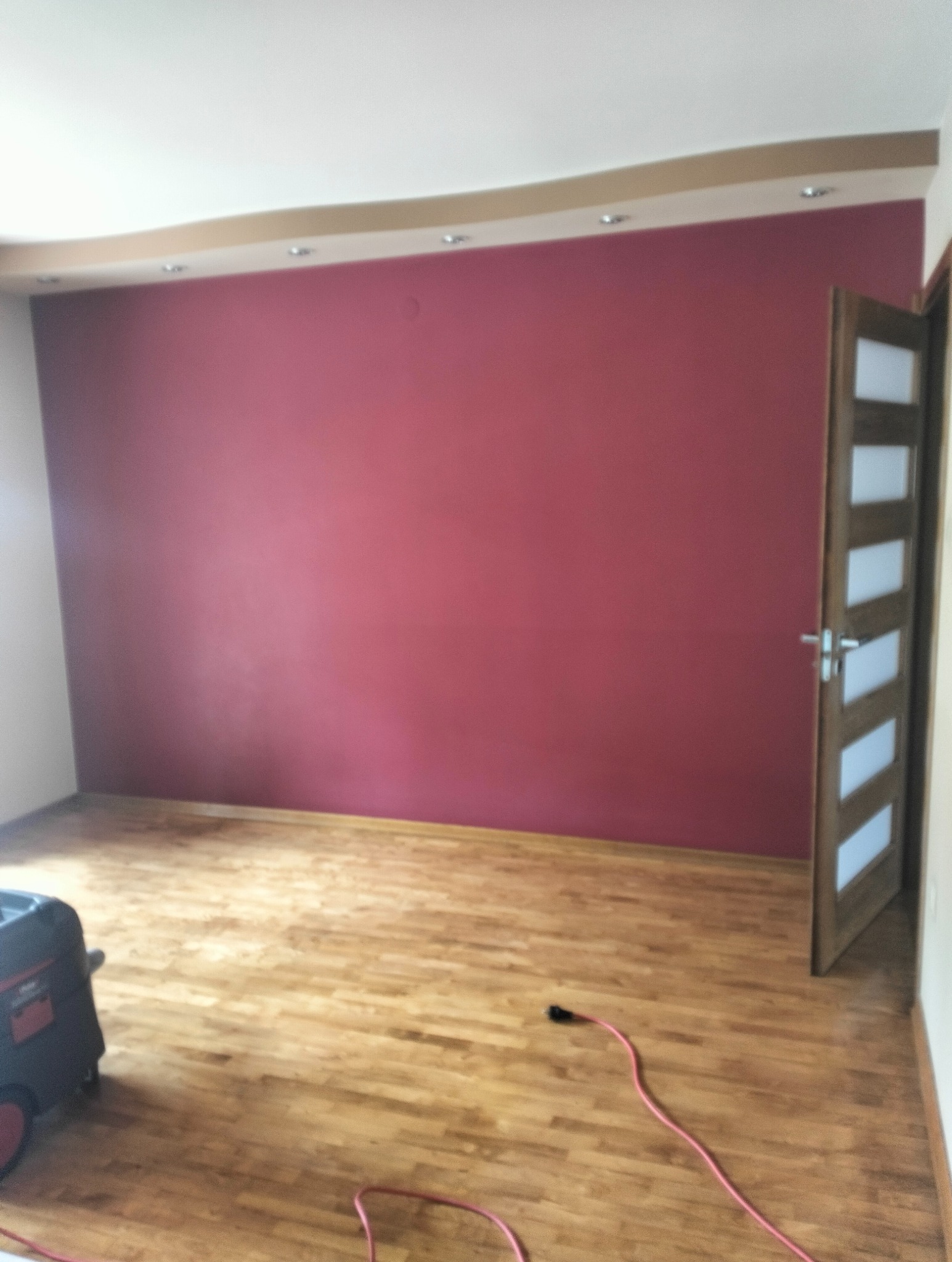 Ściana pomalowana na bordowo w pokoju z drewnianą podłogą i otwartymi drzwiami. Widoczny odkurzacz przemysłowy i kabel zasilający.