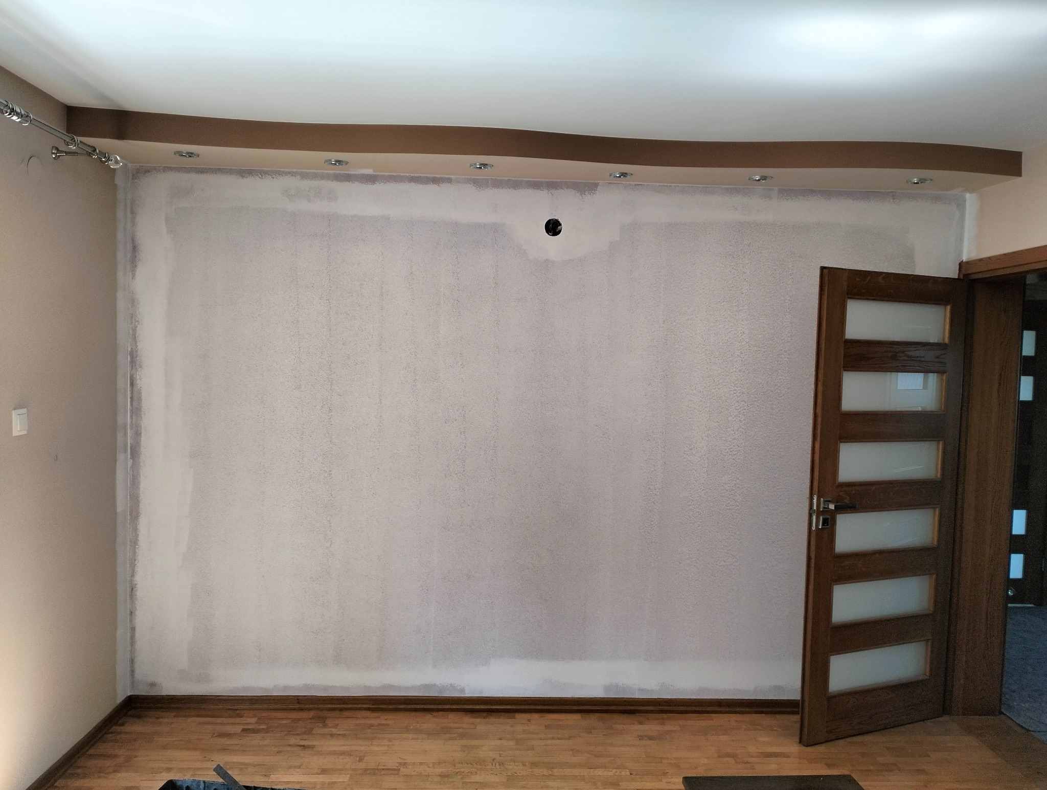 Ściana w trakcie szpachlowania, widoczne ślady po nałożeniu gładzi. Drewniana podłoga, drzwi z przeszkleniami po prawej. Sufit z oświetleniem punktowym.