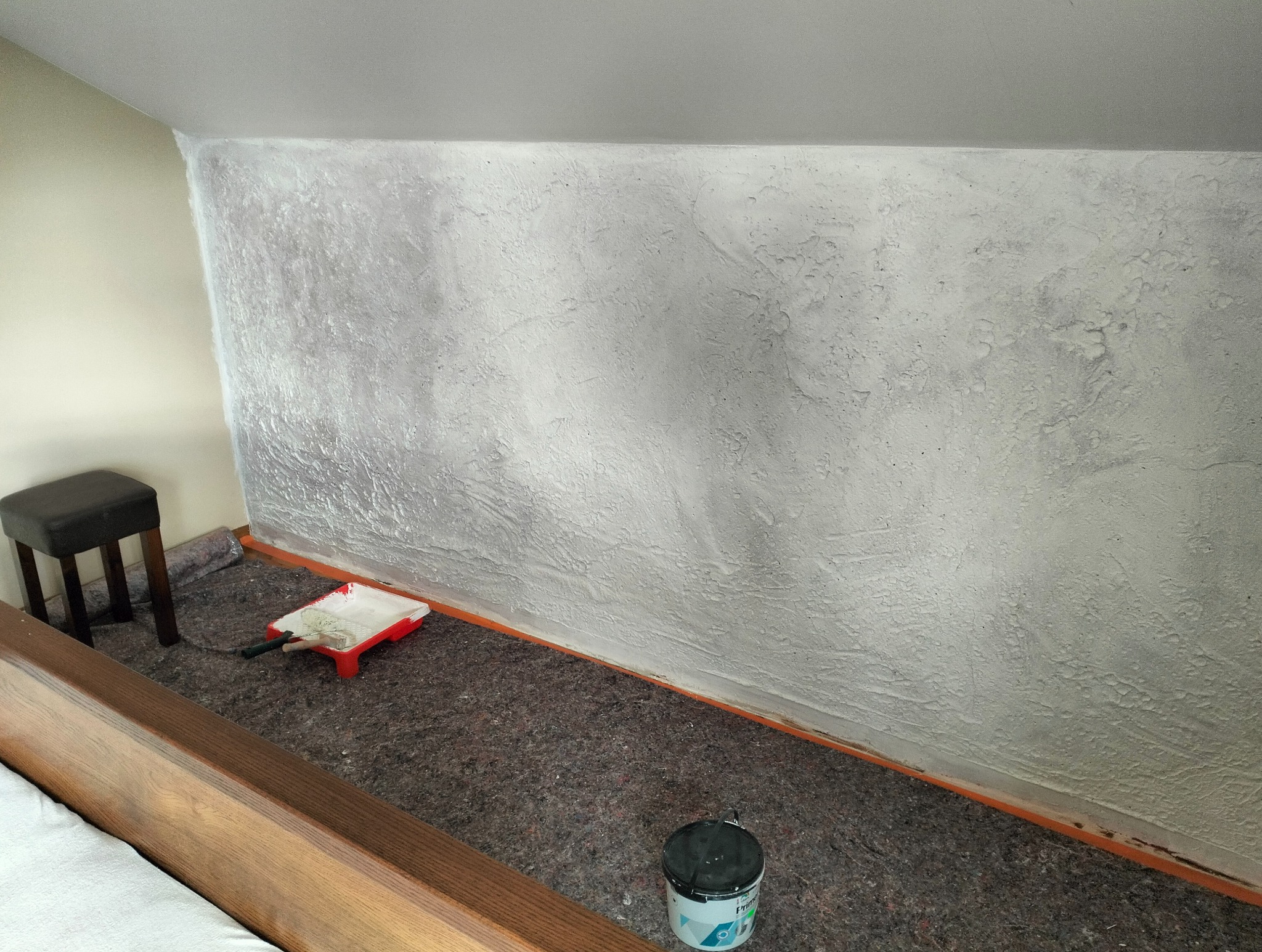 Teksturowana ściana w odcieniach szarości w trakcie malowania, zabezpieczona taśmą. Na podłodze kuweta malarska i pędzel, widoczny fragment mebla i stołek.