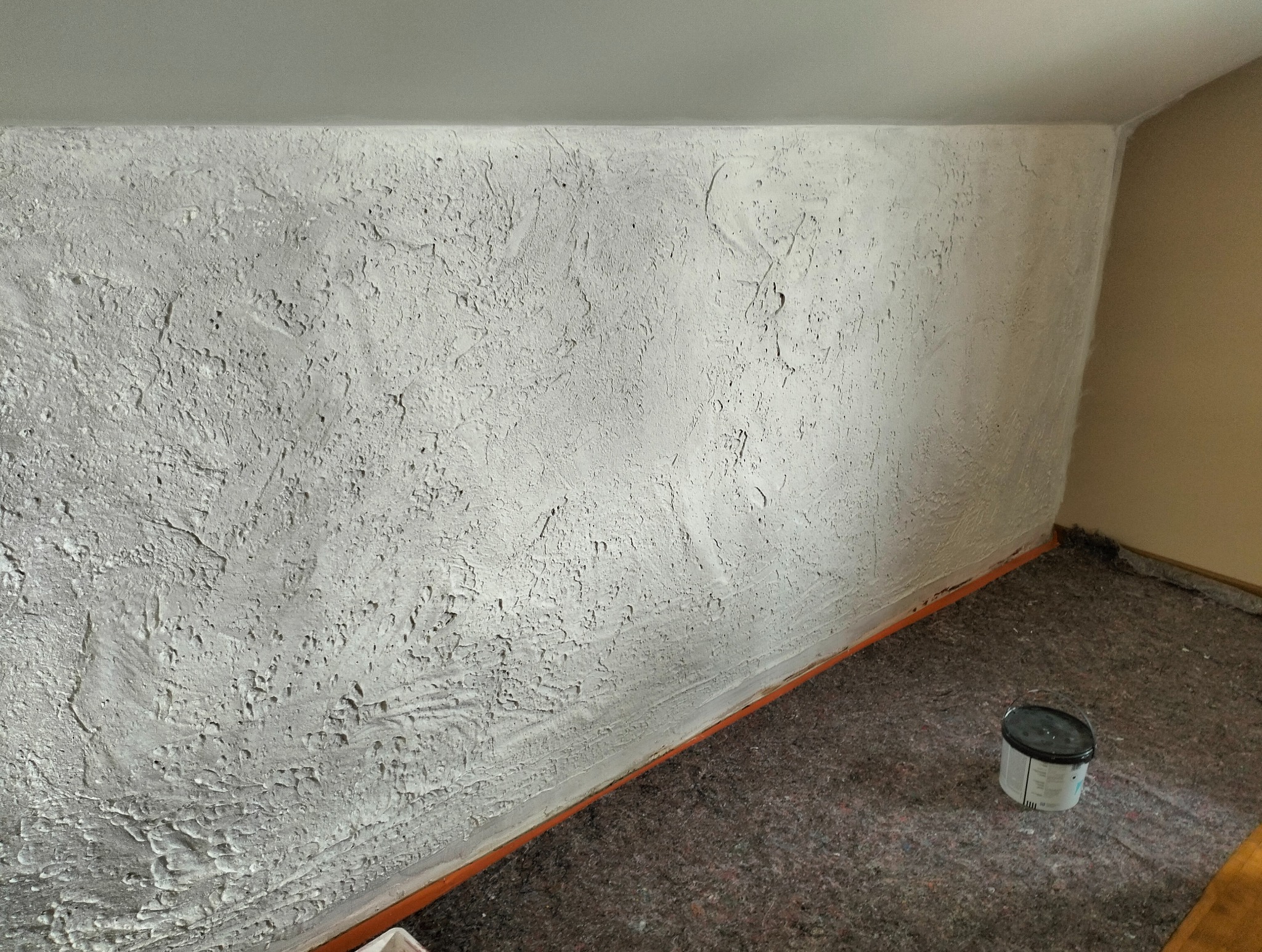 Teksturowana, biała ściana po szpachlowaniu w skosie poddasza, zabezpieczona folią i z wiaderkiem farby. Widok na wykończone wnętrze.