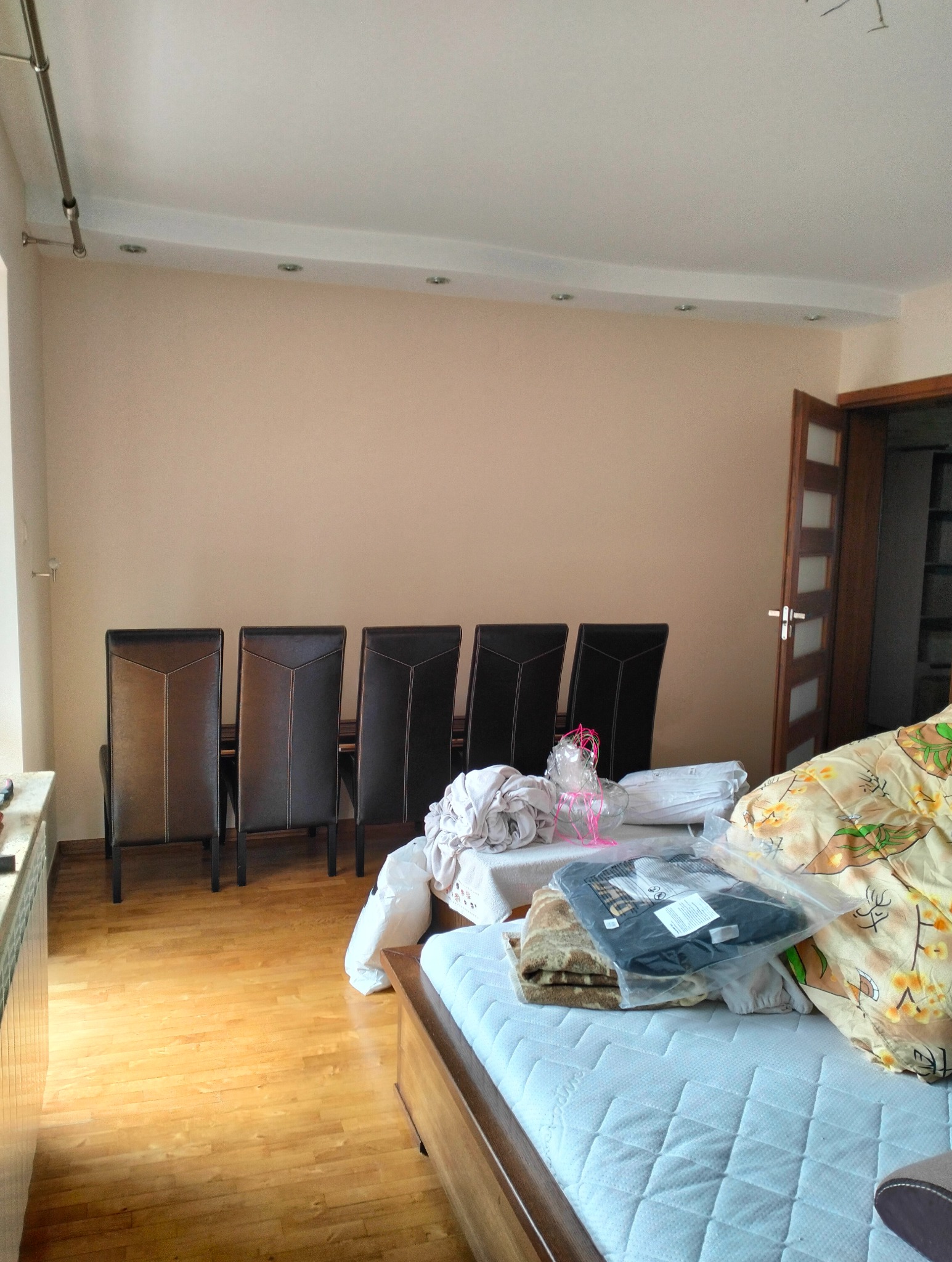 Jasne wnętrze pokoju z parkietem, kremową ścianą i rzędem krzeseł. Na łóżku leżą tekstylia i folie, sugerując przygotowania do remontu lub przeprowadzkę.