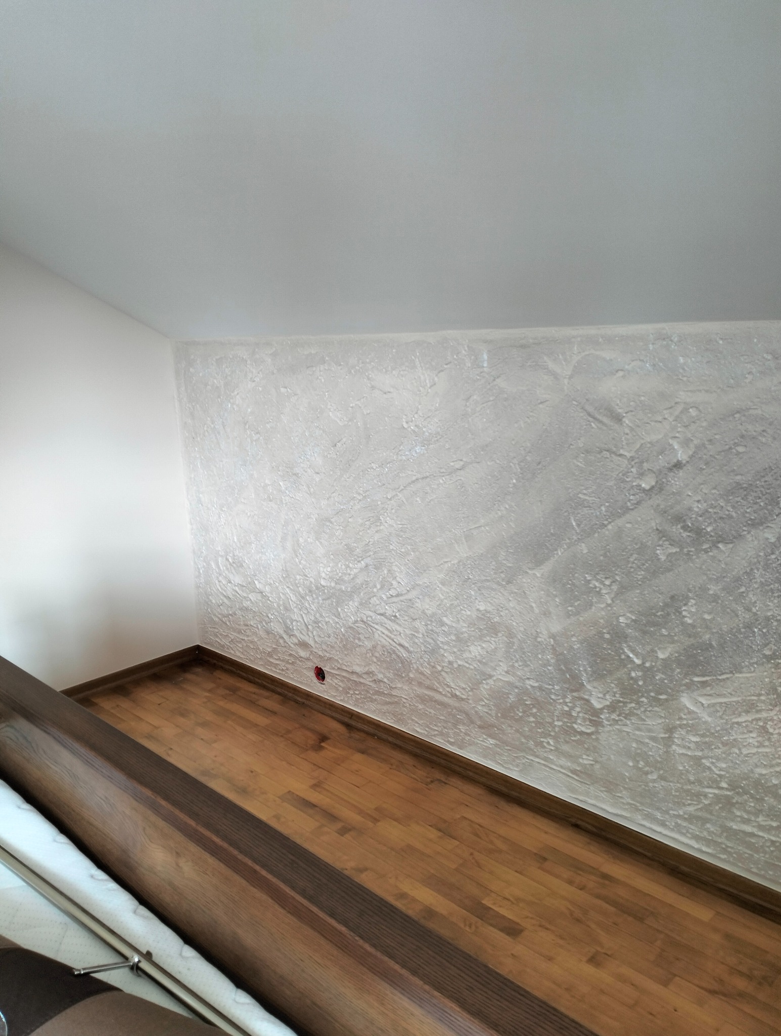 Wykończone wnętrze: drewniana podłoga, ściana z dekoracyjną, teksturowaną szpachlą w odcieniach bieli i szarości, widoczny fragment balustrady. Detal gniazdka elektrycznego.
