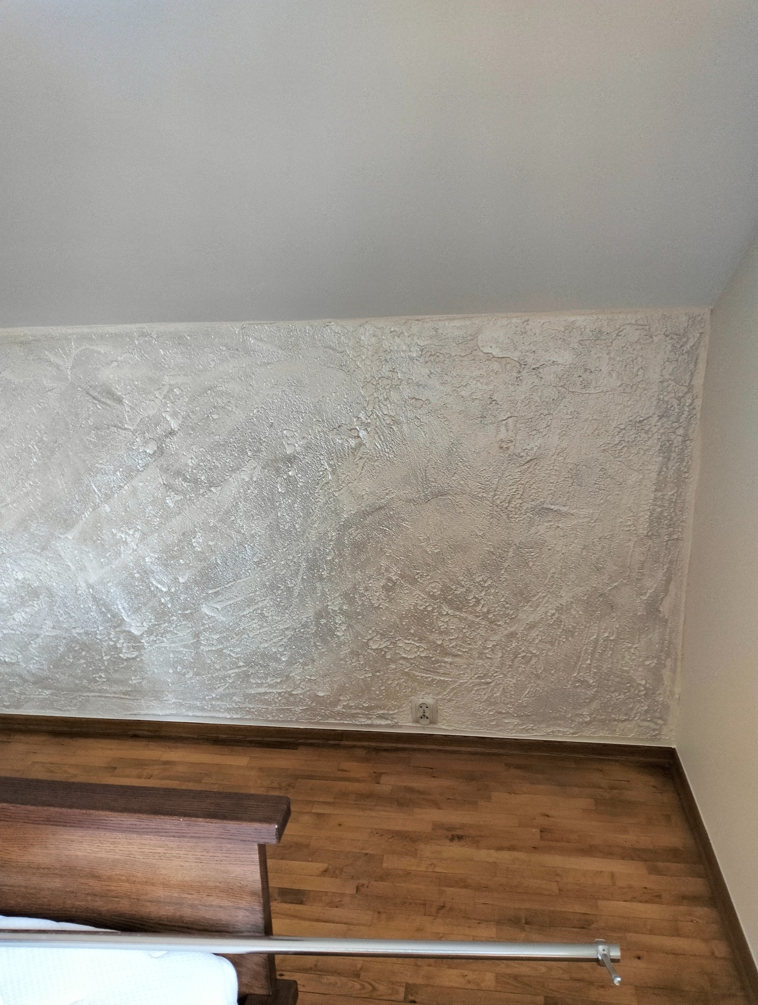 Ściana z dekoracyjną, srebrzystą fakturą w sypialni, widoczna nad drewnianą podłogą i fragmentem łóżka. Delikatne światło podkreśla unikalny wzór.