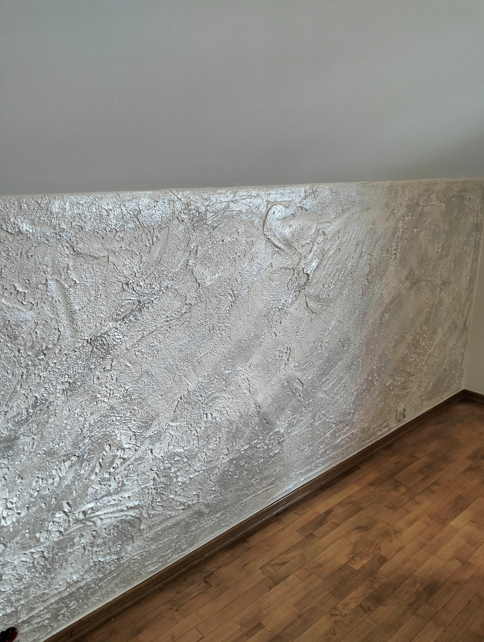 Teksturowana ściana w odcieniach szarości z efektem połysku, wykończona listwą przypodłogową i panelami podłogowymi. Widok na fragment poddasza.