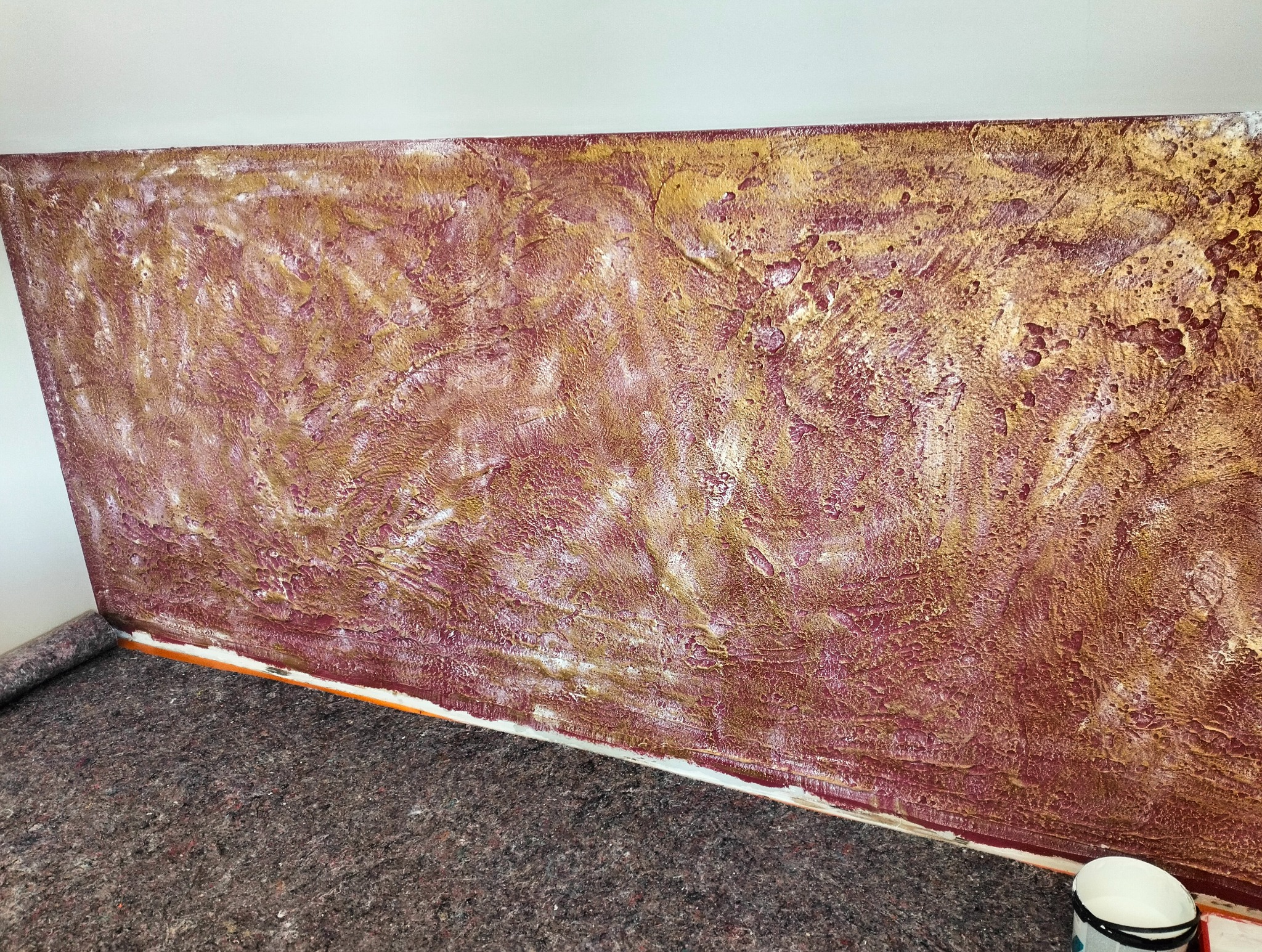 Dekoracyjna ściana z efektem strukturalnym w odcieniach bordo i złota, zabezpieczona folią malarską. Widoczny fragment podłogi z szarą wykładziną.