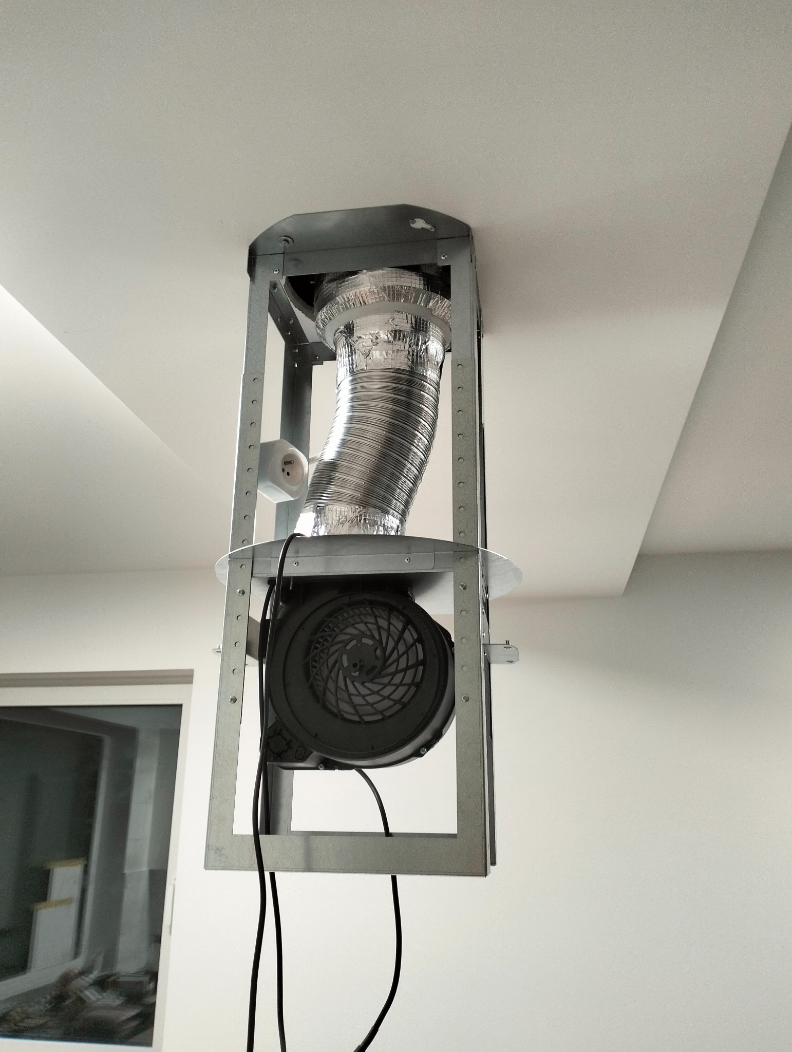 Montaż wentylacji mechanicznej w Orońsku. Widoczny wentylator w metalowej konstrukcji i rura wentylacyjna pod sufitem. Gniazdko elektryczne obok.