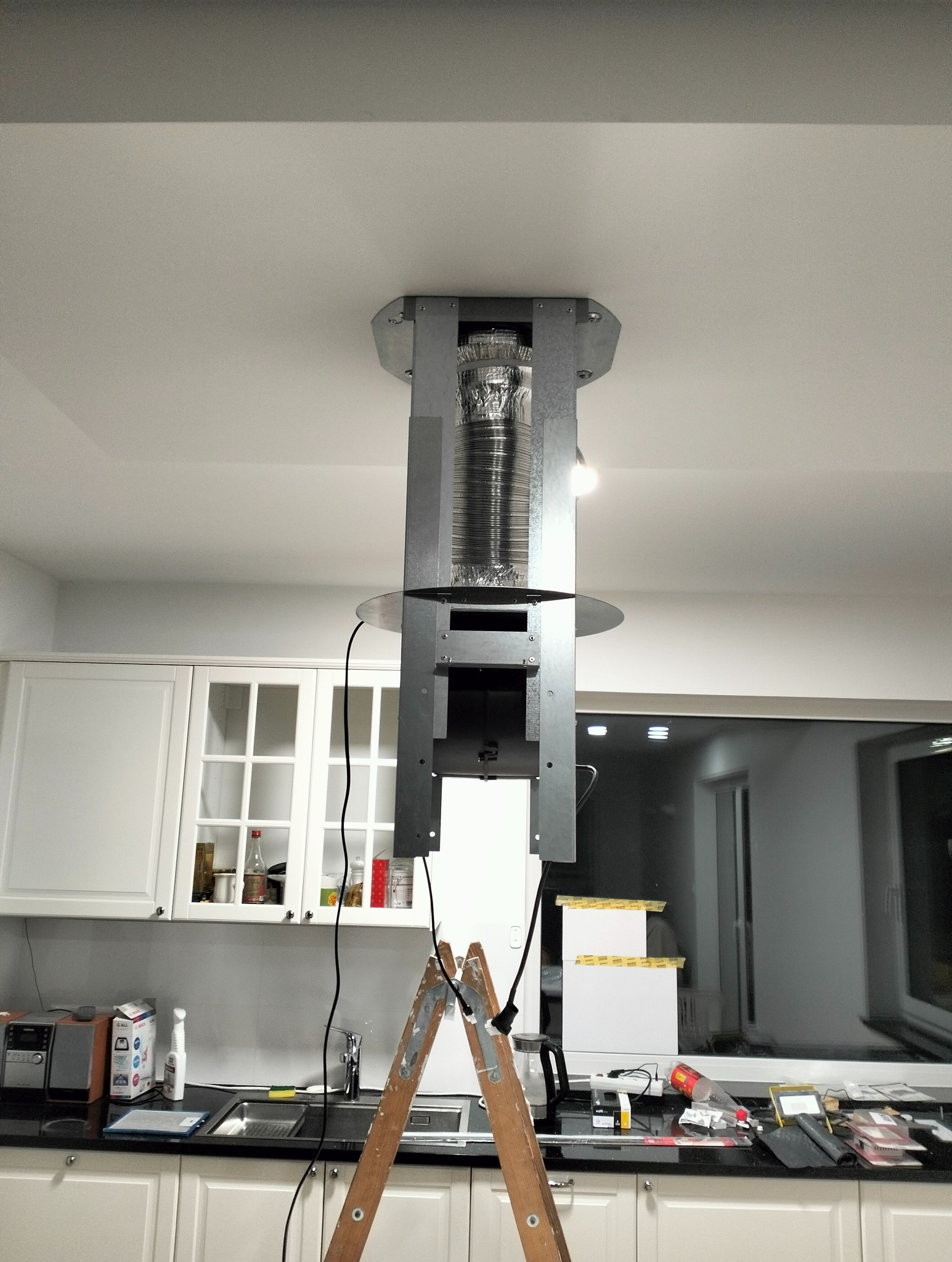 Montaż okapu kuchennego z metalową rurą wentylacyjną, widoczny drabina i fragment blatu kuchennego z wyposażeniem. Ujęcie z dołu.