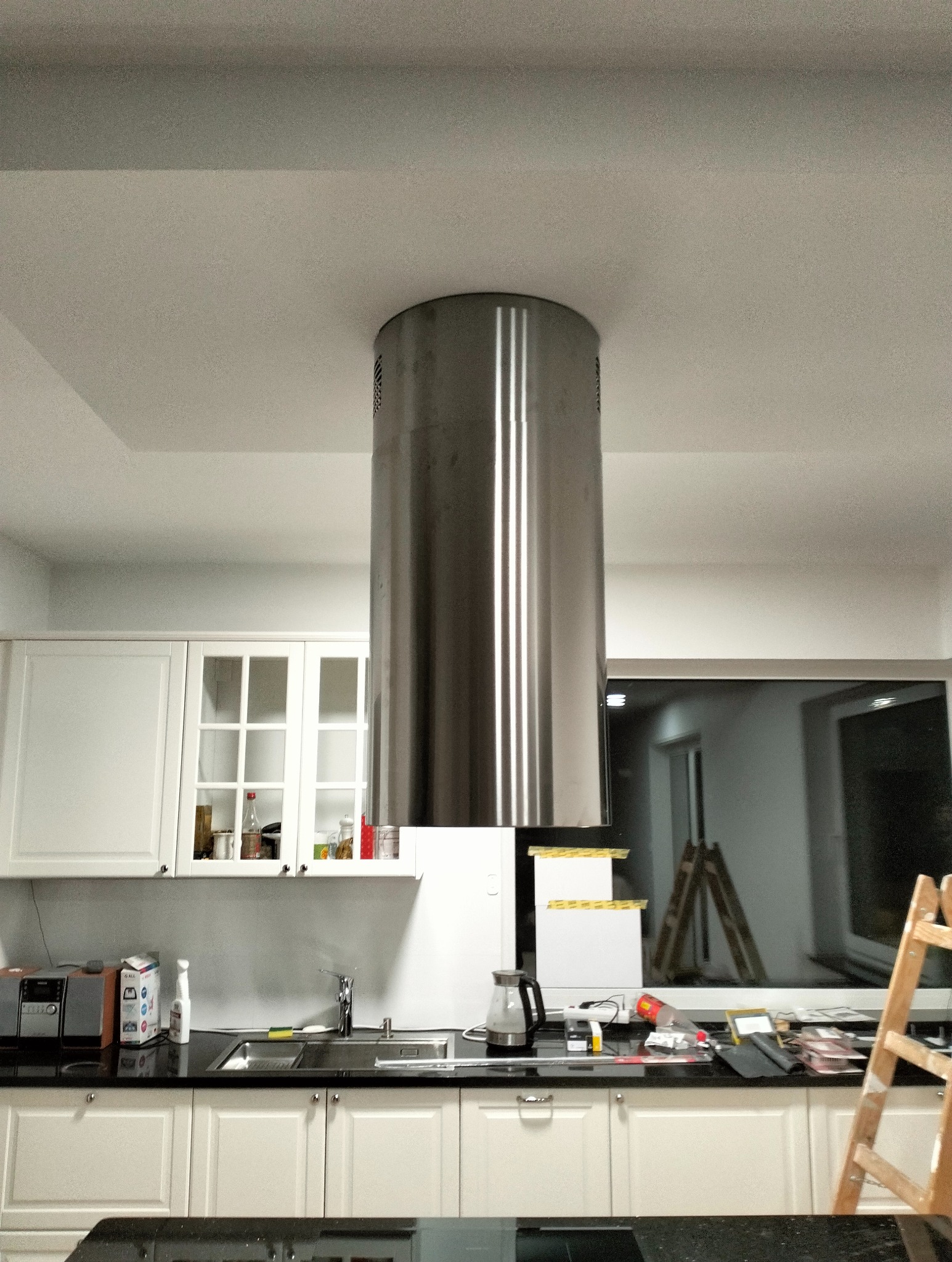 Nowoczesna kuchnia z białymi szafkami i stalowym okapem cylindrycznym, widok z dołu. W tle okno i drabina malarska.