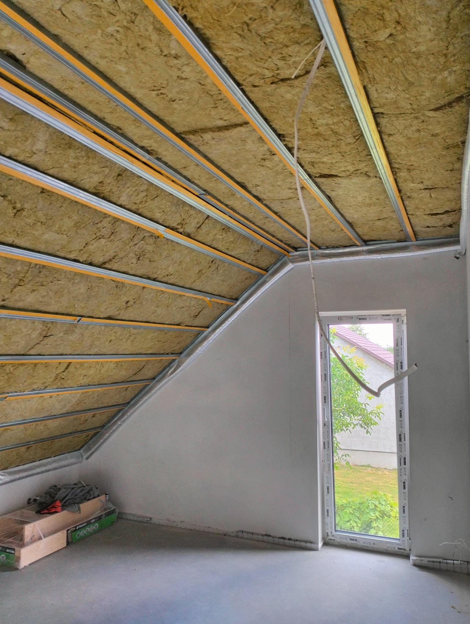 Poddasze z ociepleniem z wełny mineralnej, widoczne metalowe profile pod płyty GK. Okno z widokiem na zieleń. Etap prac wykończeniowych.