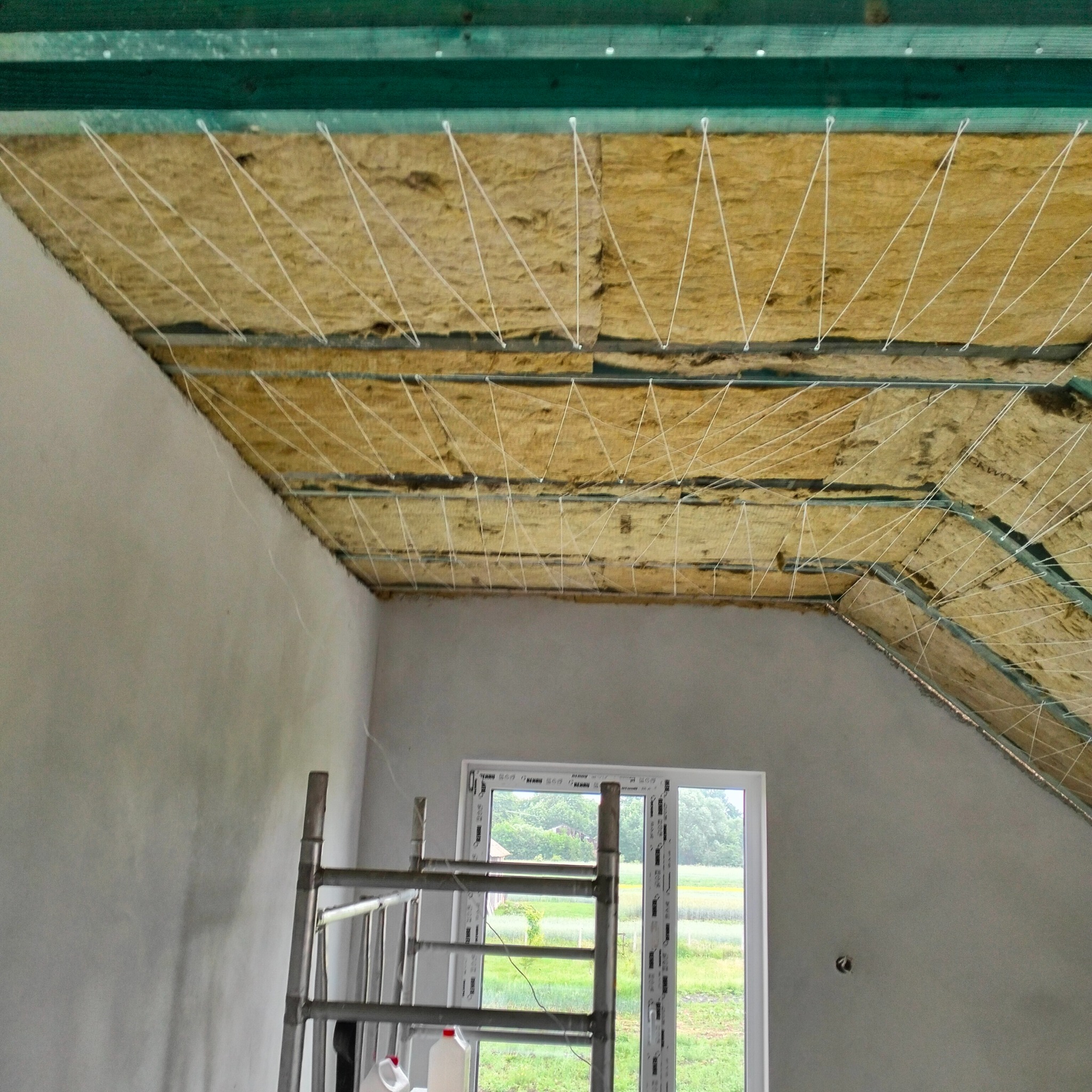 Ocieplenie poddasza wełną mineralną, widoczne sznurki mocujące. W tle okno i ściana w surowym stanie. Metalowa drabina oparta o ścianę.