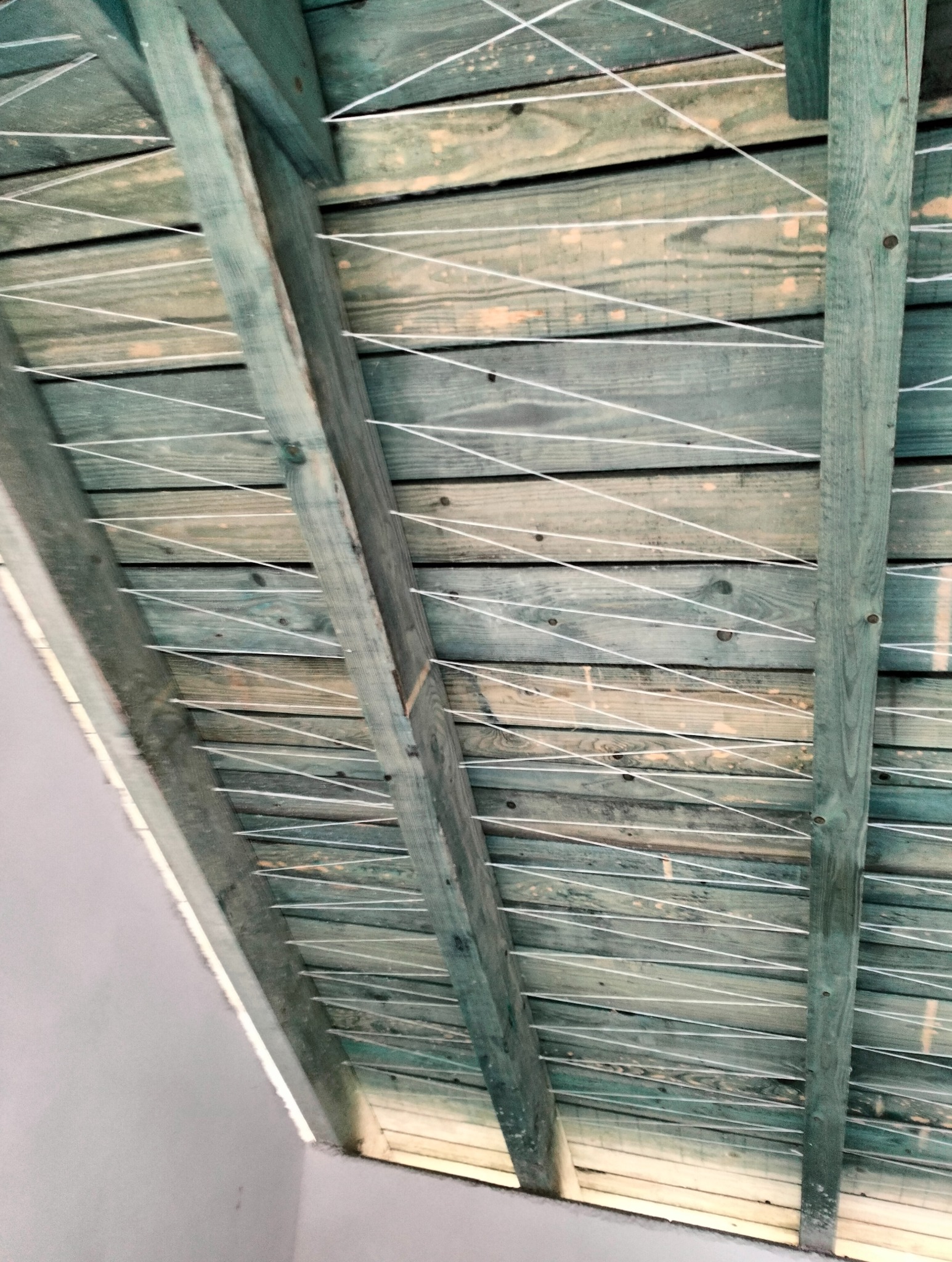 Drewniana konstrukcja poddasza z naciągniętym sznurkiem, przygotowana do ocieplenia. Widoczne deski w odcieniach zieleni i beżu oraz biały sznurek tworzący siatkę.